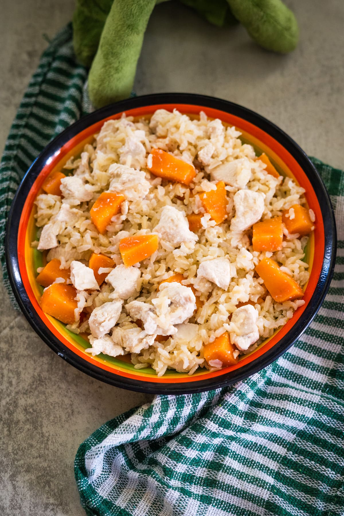 A bowl of rice mixed with chunks of chicken and pumpkin, placed on a green and white checkered cloth.