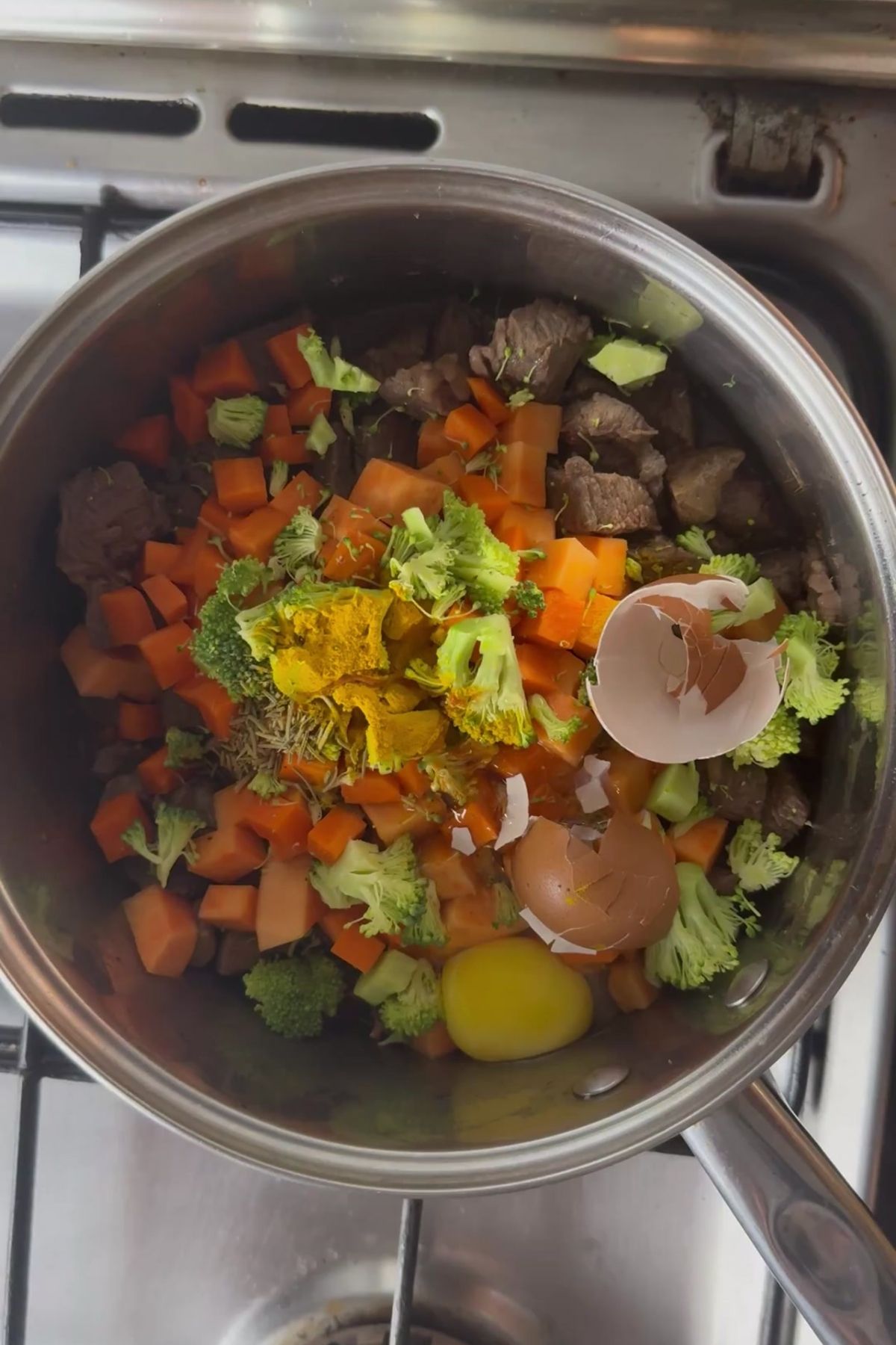 A pot on a stove contains chopped carrots, broccoli, beef pieces, spices, a whole raw egg, and broken eggshellsโperfect ingredients for a nutritious beef stew for dogs.