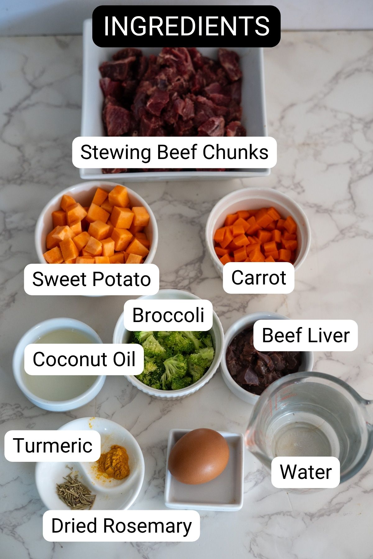 A variety of labeled ingredients on a marble surface, including stewing beef chunks, sweet potato, carrot, broccoli, beef liver, coconut oil, turmeric, dried rosemary, egg, and waterโperfect for making homemade beef stew for dogs.