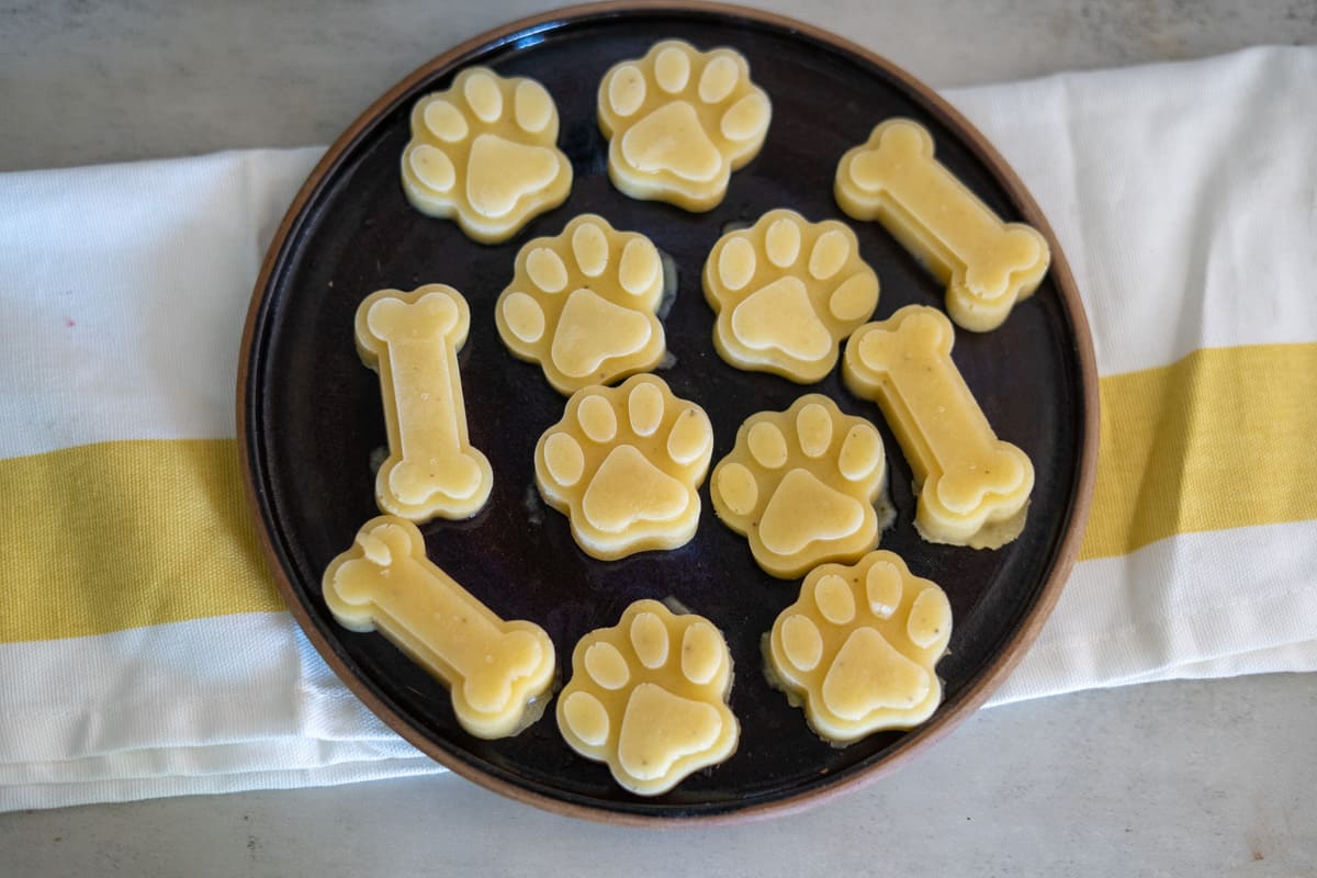 A dark plate with bone-shaped and paw-shaped banana dog treats arranged on top of a white and yellow cloth.