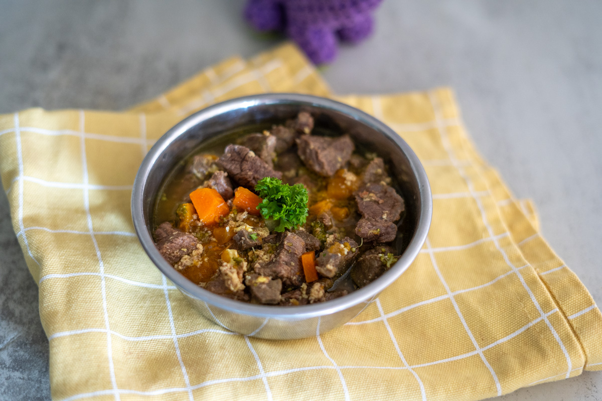A metal bowl filled with beef stew for dogs, featuring chunks of meat, carrots, and other vegetables, garnished with parsley, sits on a yellow checkered cloth.