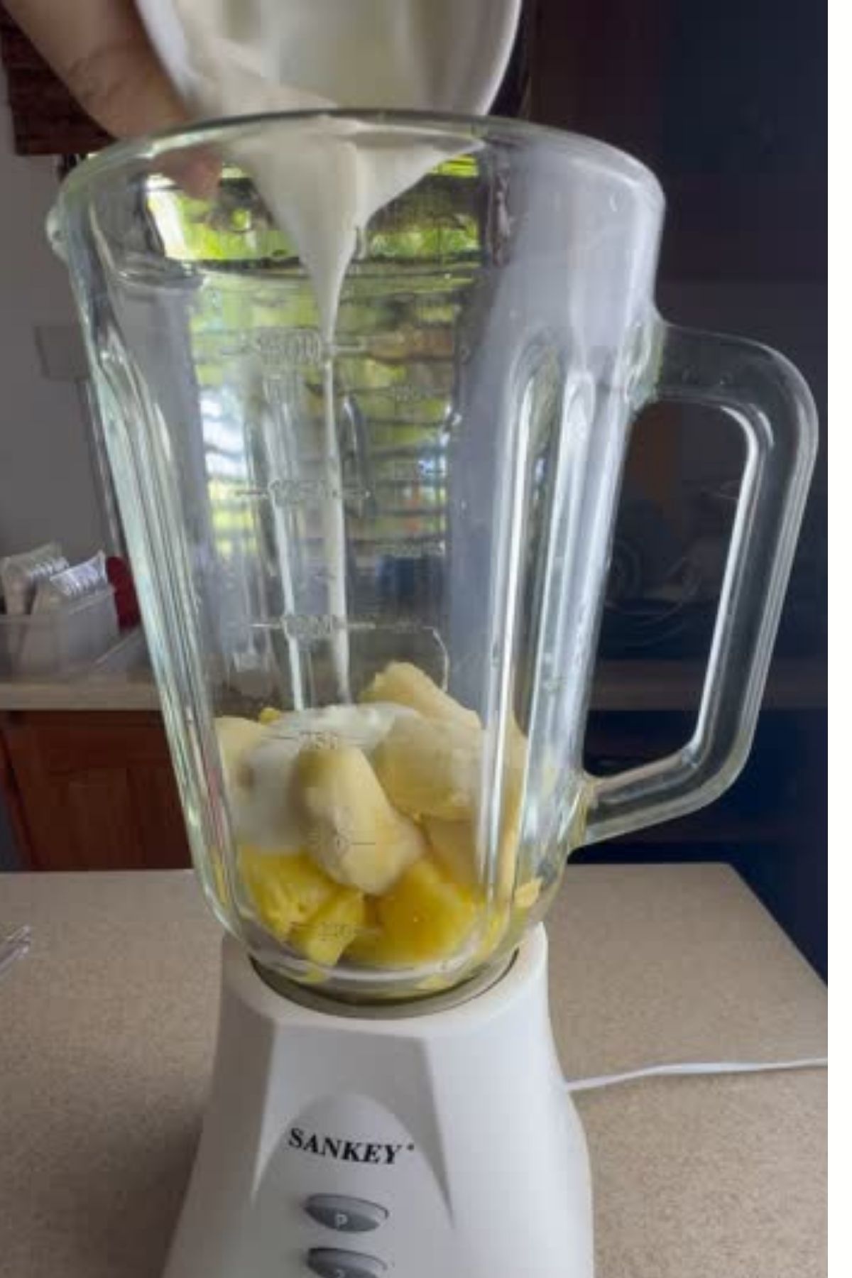 A blender with chopped fruit, perfect for making banana dog treats, is being filled with a white liquid, likely milk or yogurt, from a cup.