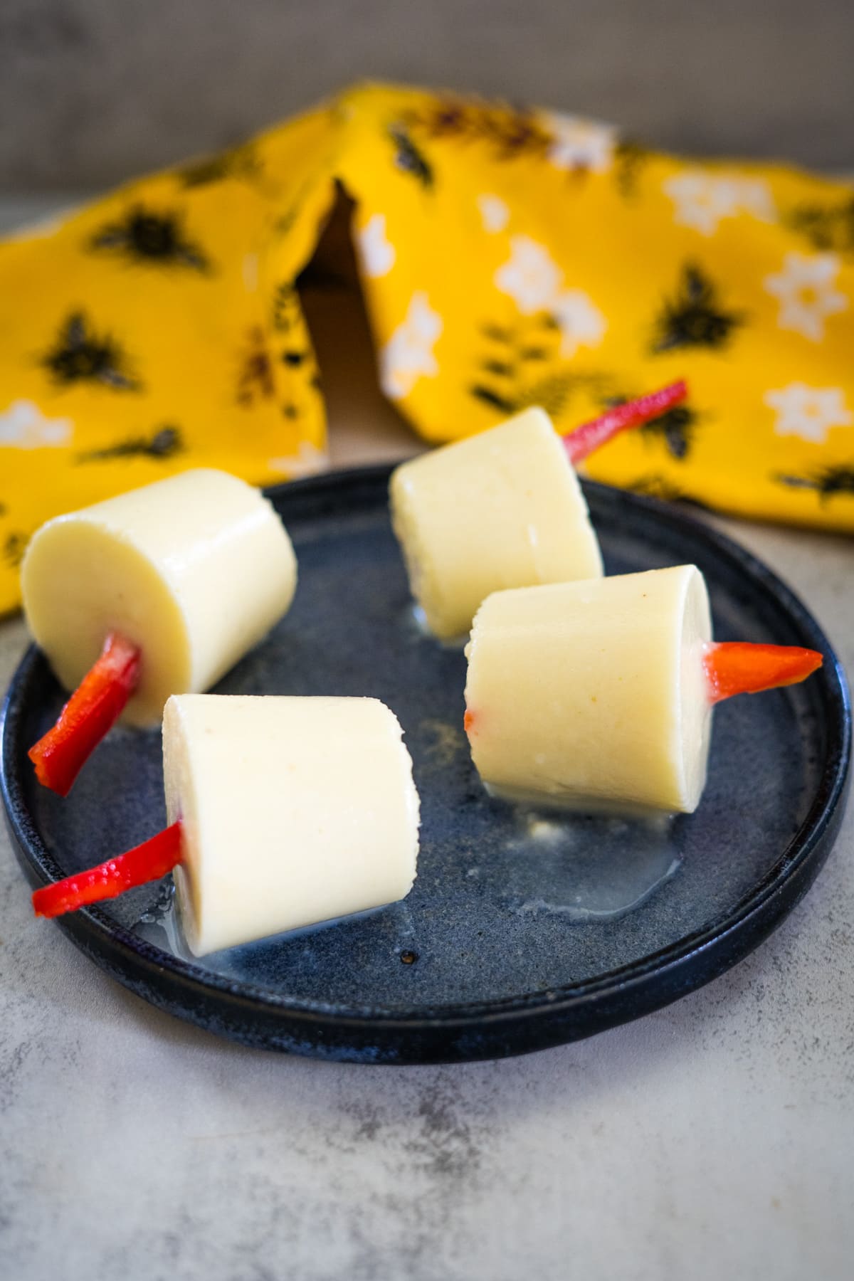 Four creamy pupsicles with red sticks rest on a dark plate, complemented by a yellow floral cloth in the background.