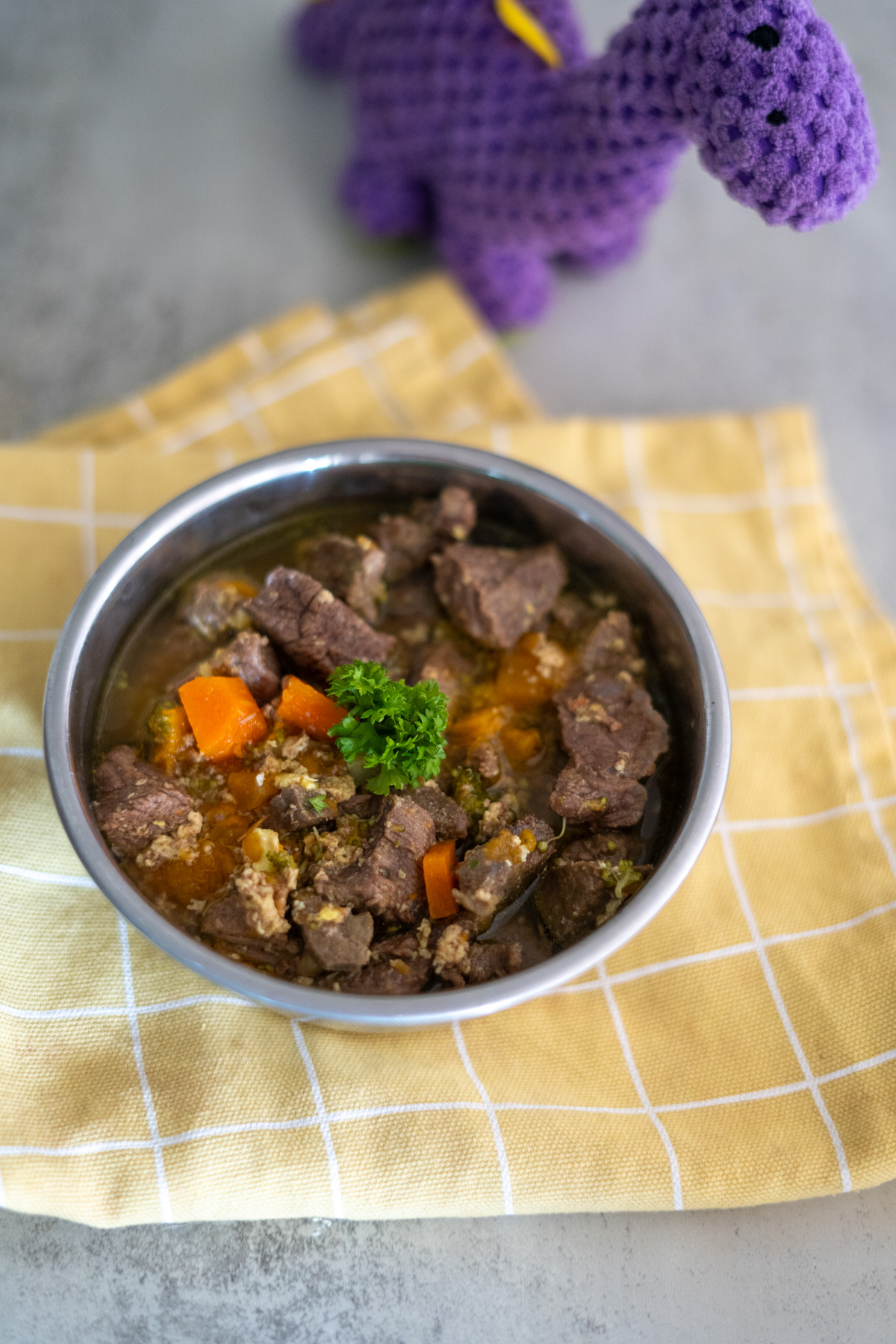 A metal bowl filled with beef stew for dogs, containing chunks of meat and carrots, sits on a yellow checkered cloth, with a purple crocheted dinosaur toy in the background.
