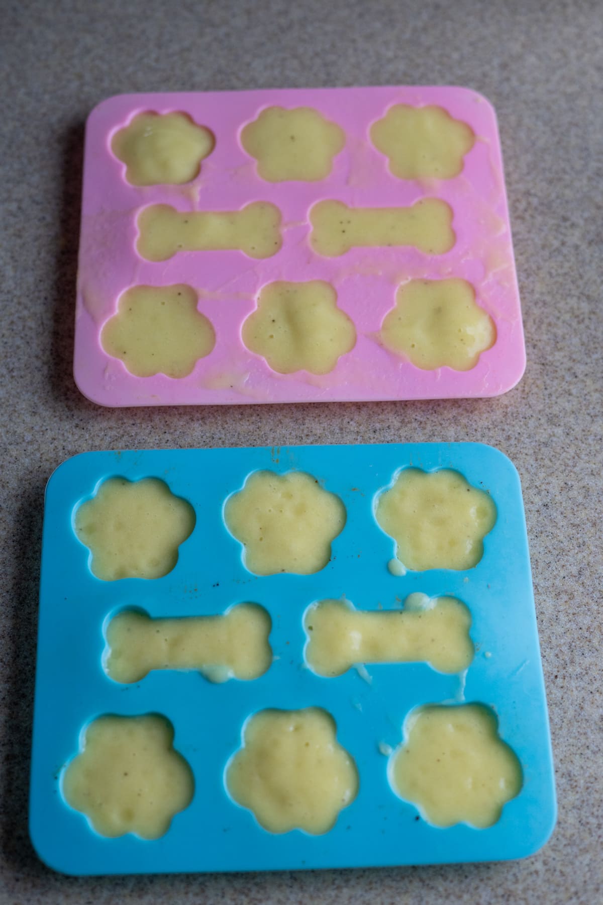 Two silicone molds, one pink and one blue, are filled with banana dog treats batter; each mold features spaces shaped like bones and paw prints.