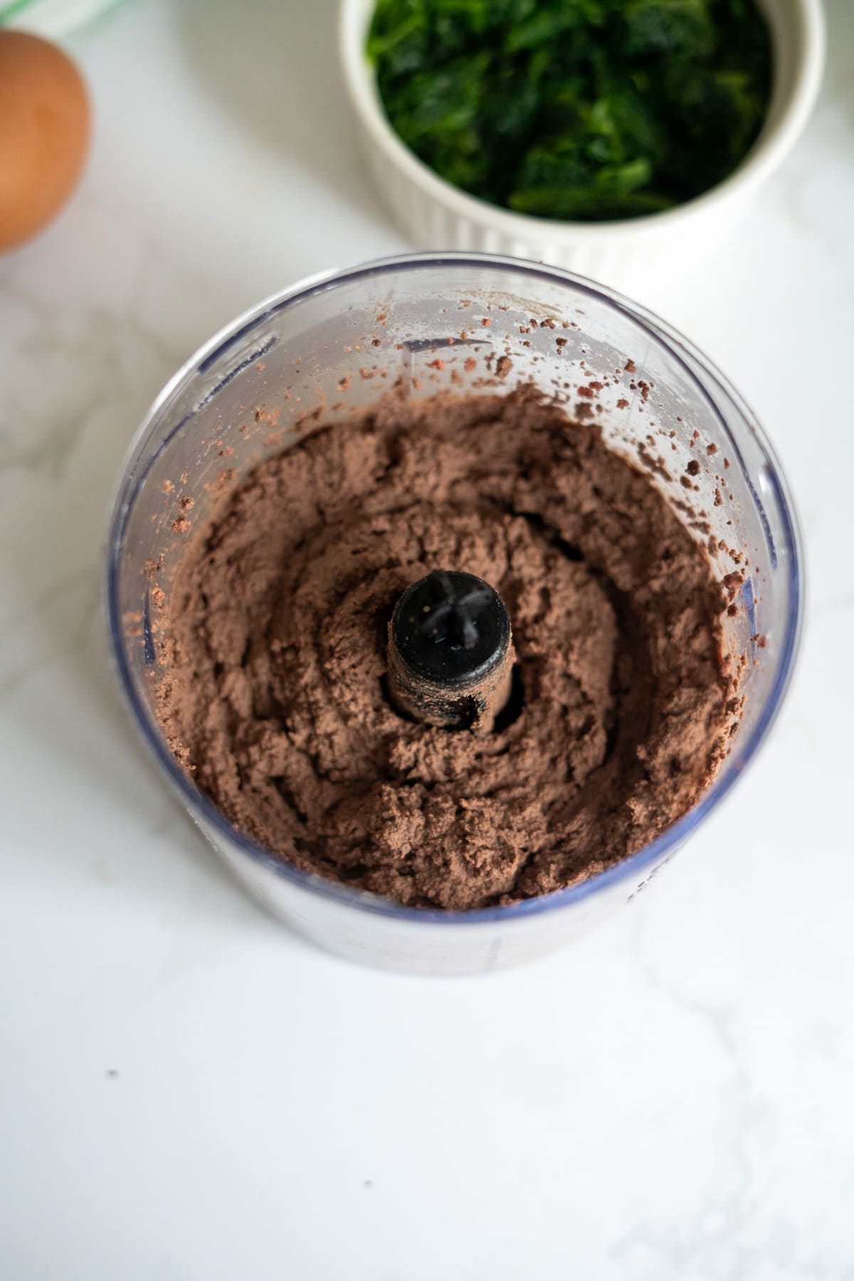 A food processor contains a thick chocolate mixture. In the background, there is a white ramekin with green leafy contents and part of a brown egg is visible—ingredients often used in chicken liver meatballs for dogs.