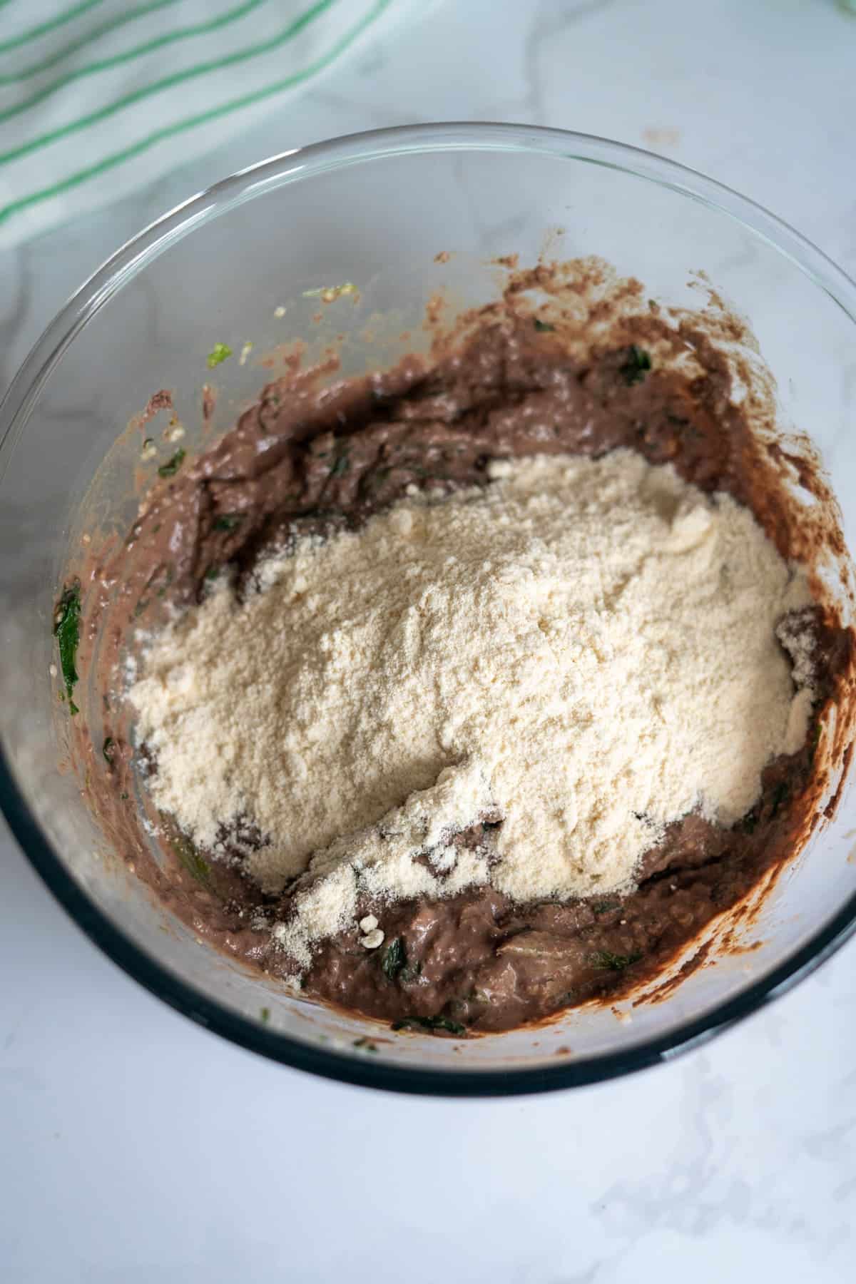 A glass mixing bowl containing a dark, chunky mixture—perfect for chicken liver meatballs for dogs—with a layer of light-coloured flour or powder on top, sitting on a white surface.