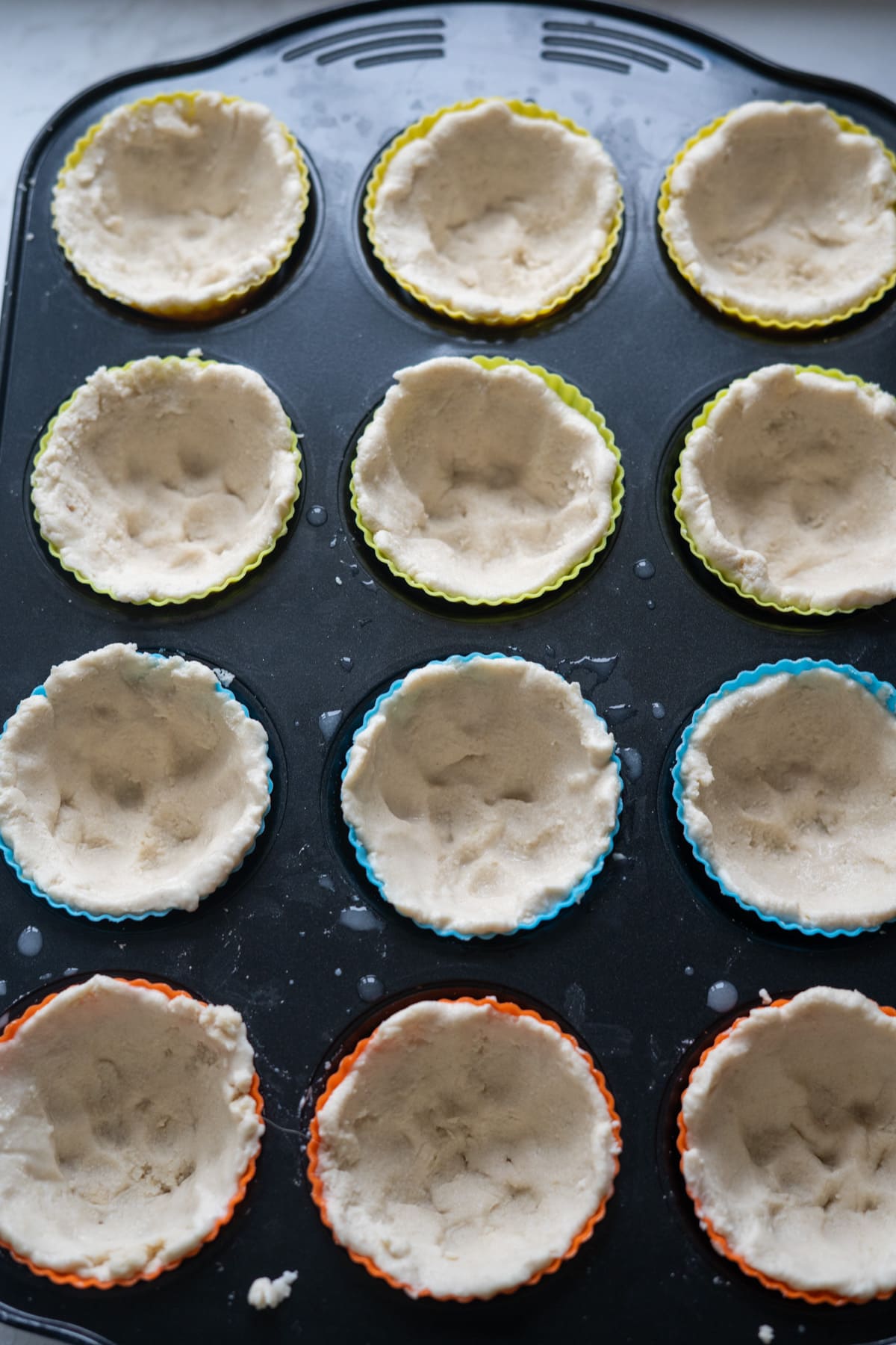 A muffin tin filled with colorful silicone liners, each holding an unbaked pie crust pressed into shape, ready to be transformed into pumpkin pies for dogs.