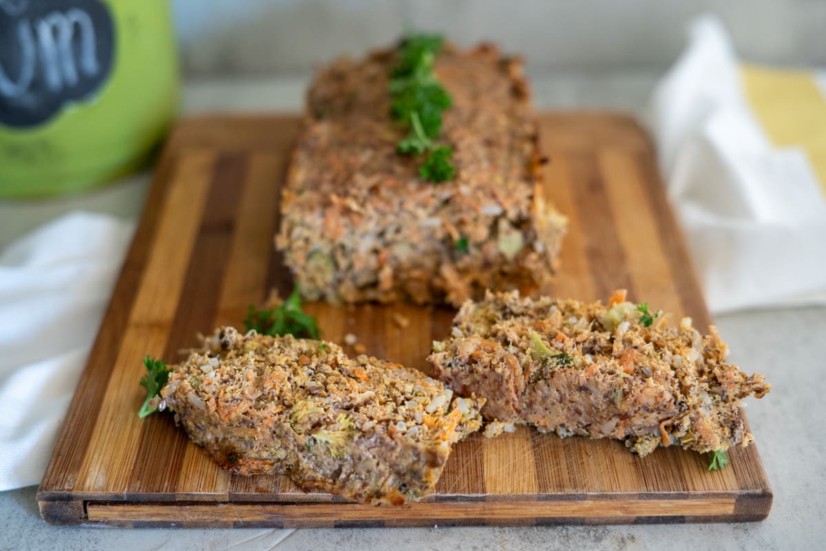 A loaf of vegetable and nut roast sits elegantly on a wooden board, reminiscent of a turkey meatloaf for dogs, with two slices perfectly cut and garnished with fresh herbs.