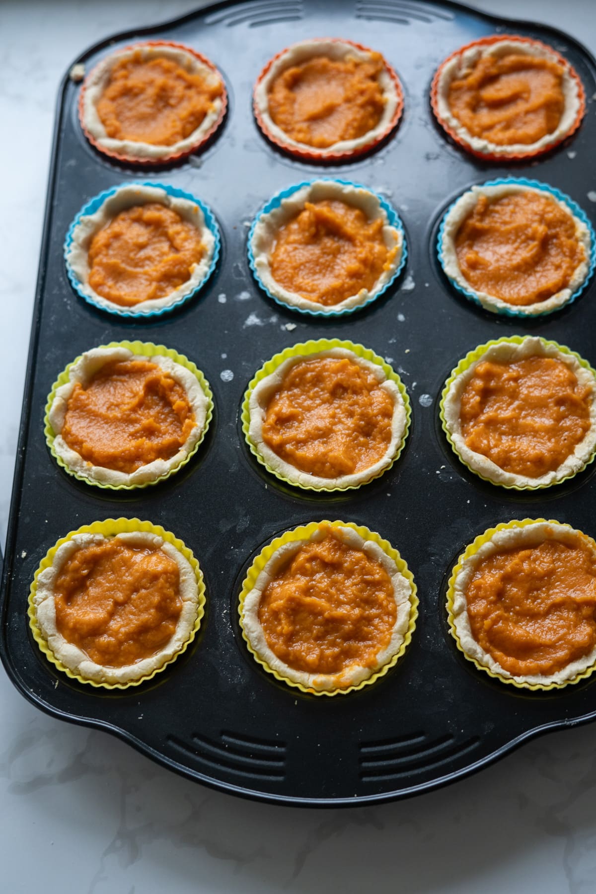 A muffin tin holds twelve unbaked mini pumpkin pies for dogs with orange filling, each lined with colorful silicone baking cups.