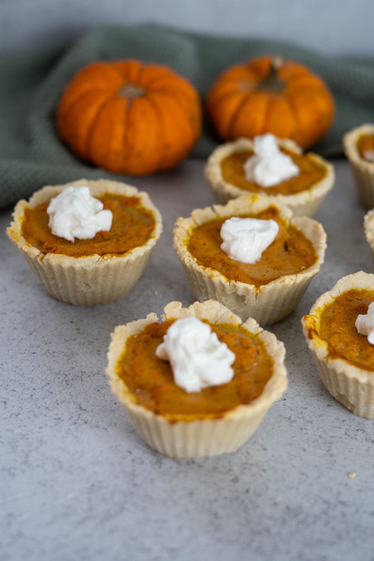 Mini pumpkin pies for dogs with whipped cream topping are arranged on a gray surface, with two small pumpkins and a green cloth in the background.
