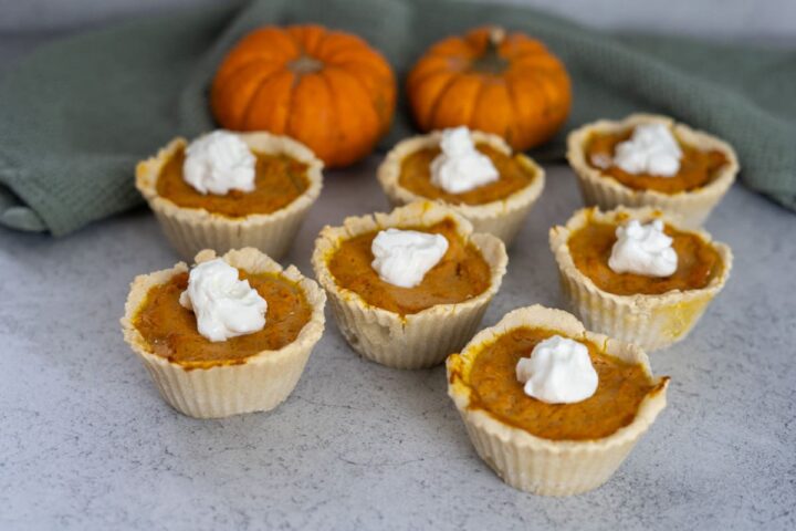 Eight mini pumpkin pies for dogs topped with whipped cream are arranged on a countertop, with two small pumpkins and a green cloth in the background.
