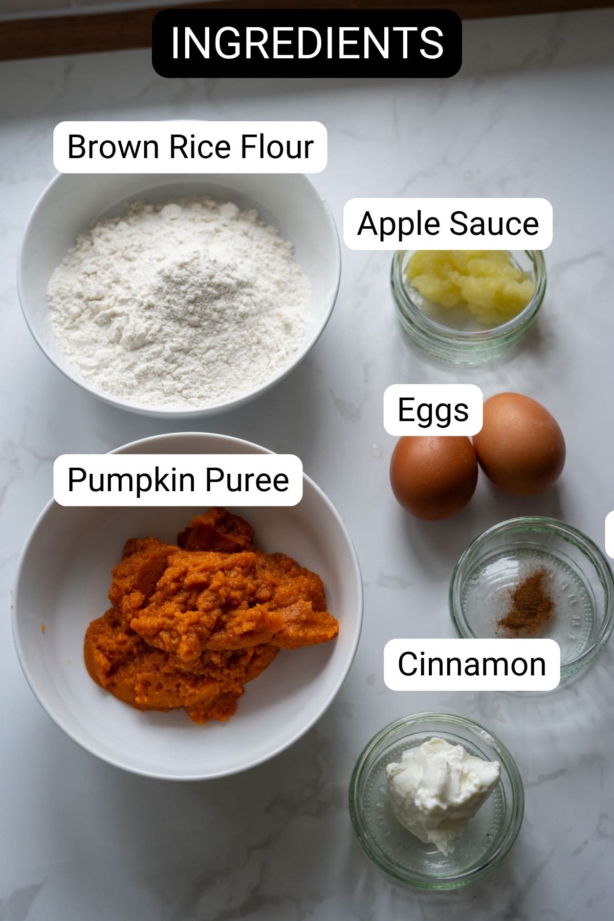 Bowls and jars containing brown rice flour, apple sauce, eggs, pumpkin puree, cinnamon, and a white ingredient are labeled on a white surface—perfect for making pumpkin pies for dogs. "Ingredients" is written at the top.