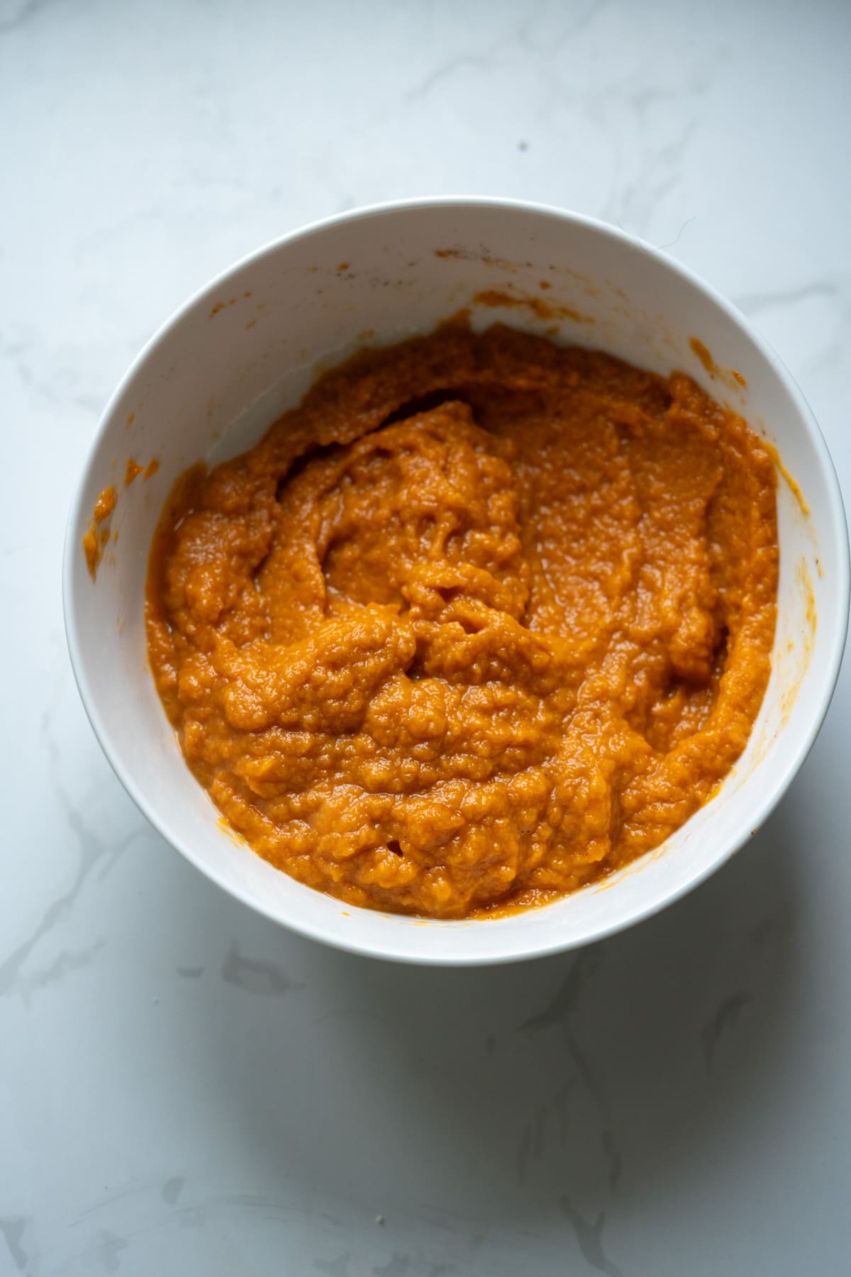 A white bowl filled with an orange sweet potato puree, perfect as a base for pumpkin pies for dogs, sits on a light marble surface.