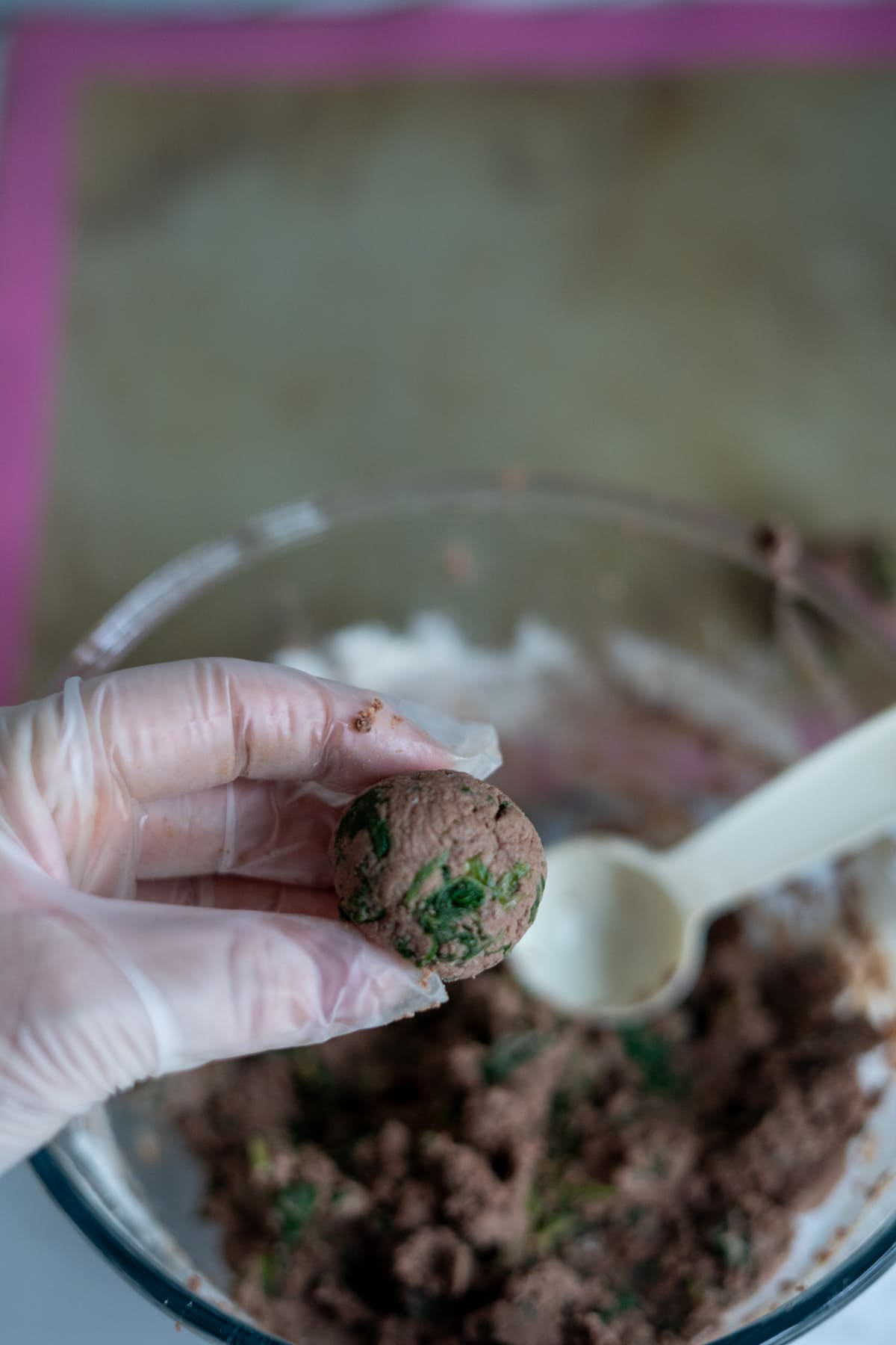 A gloved hand holding a small, round mixture of chopped greens and other ingredients—perfect chicken liver meatballs for dogs—over a bowl of similar mixture with a white spoon inside.