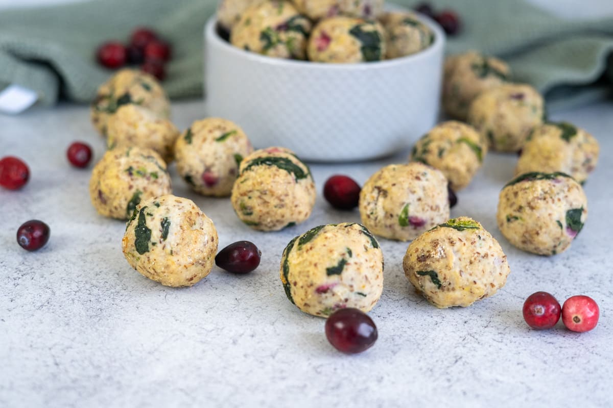 Round stuffing balls with visible herbs and cranberries are arranged on a countertop, resembling a festive meatballs for dogs recipe, with loose cranberries scattered around and a bowl of more stuffing balls in the background.