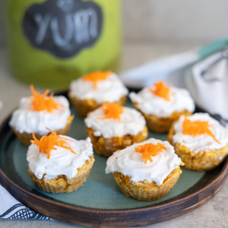 Seven carrot pupcakes topped with white frosting and shredded carrot are arranged on a round blue plate. A green container labeled "YUM" is blurred in the background.