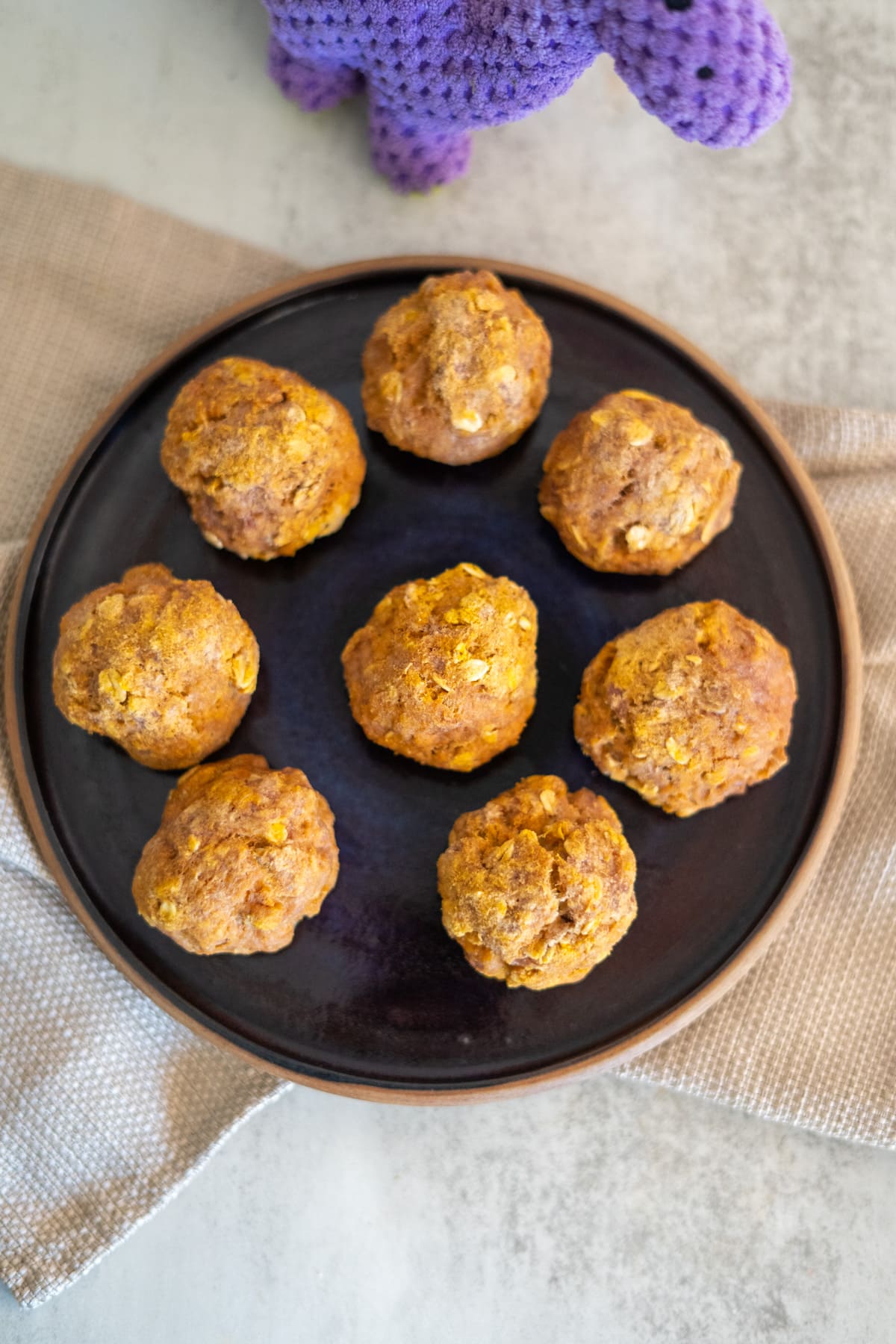 Seven golden-brown muffins on a dark plate, like delectable turkey pumpkin treats, with textured purple and beige cloth nearby.