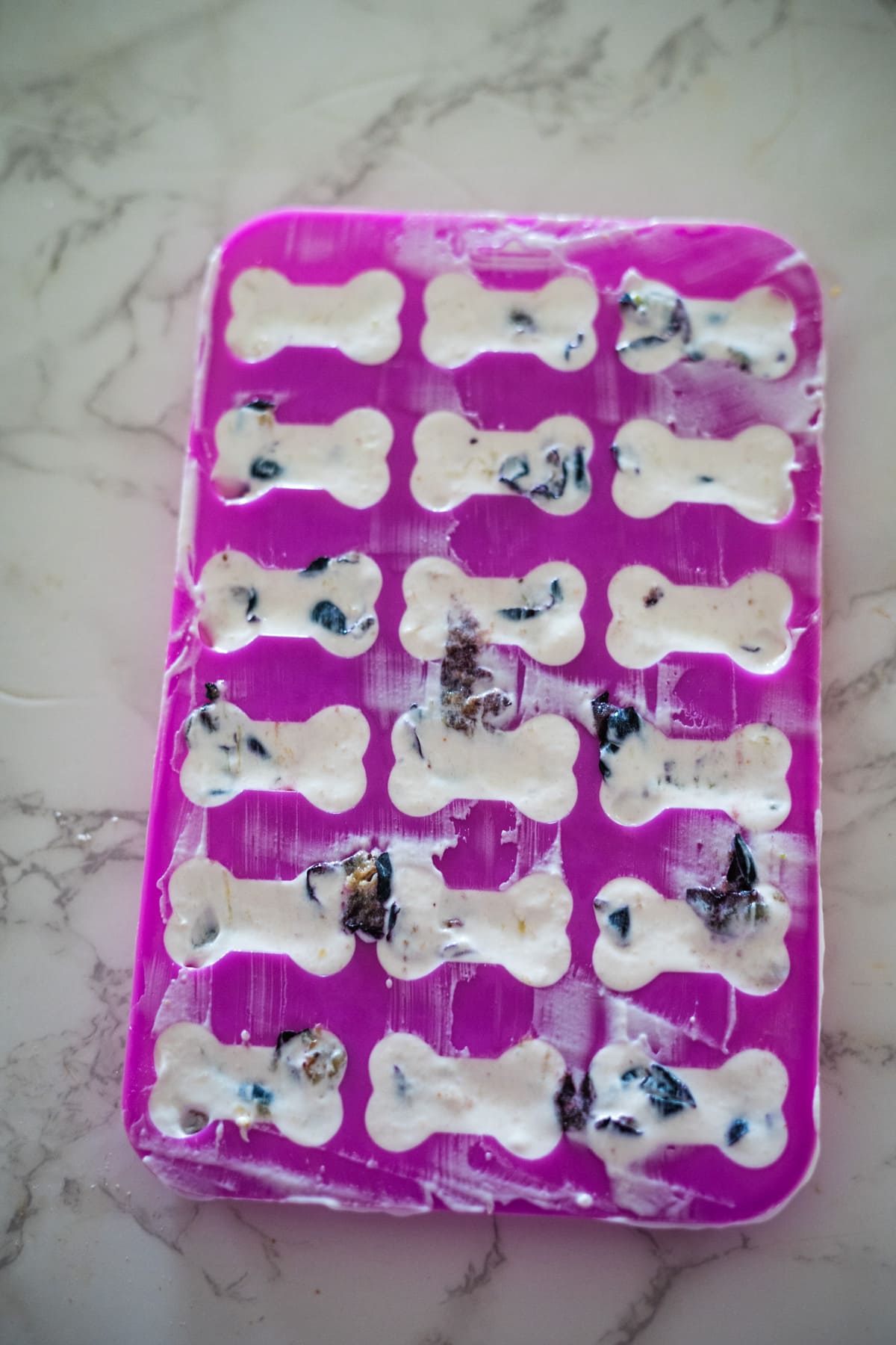 Purple silicone mold filled with blueberry yogurt dog treats, shaped like bones and partially mixed with dark berries, on a marble surface.