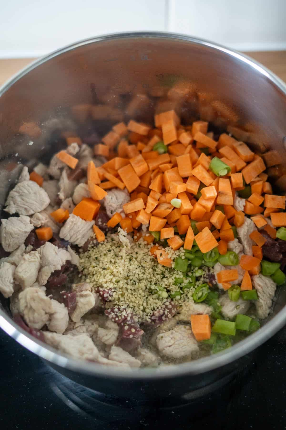 A stainless steel saucepan containing chopped raw carrots, celery, chunks of meat, and a sprinkle of seeds or grains on a hob—perfect for preparing a wholesome turkey dog food recipe.