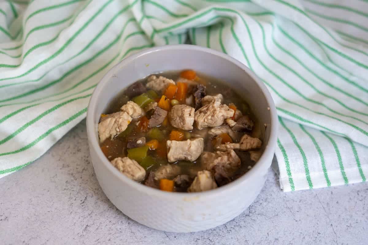 A white bowl filled with a chunky soup, similar to a turkey dog food recipe, containing pieces of meat, carrots, celery, and broth sits on a light surface next to a green and white striped cloth.