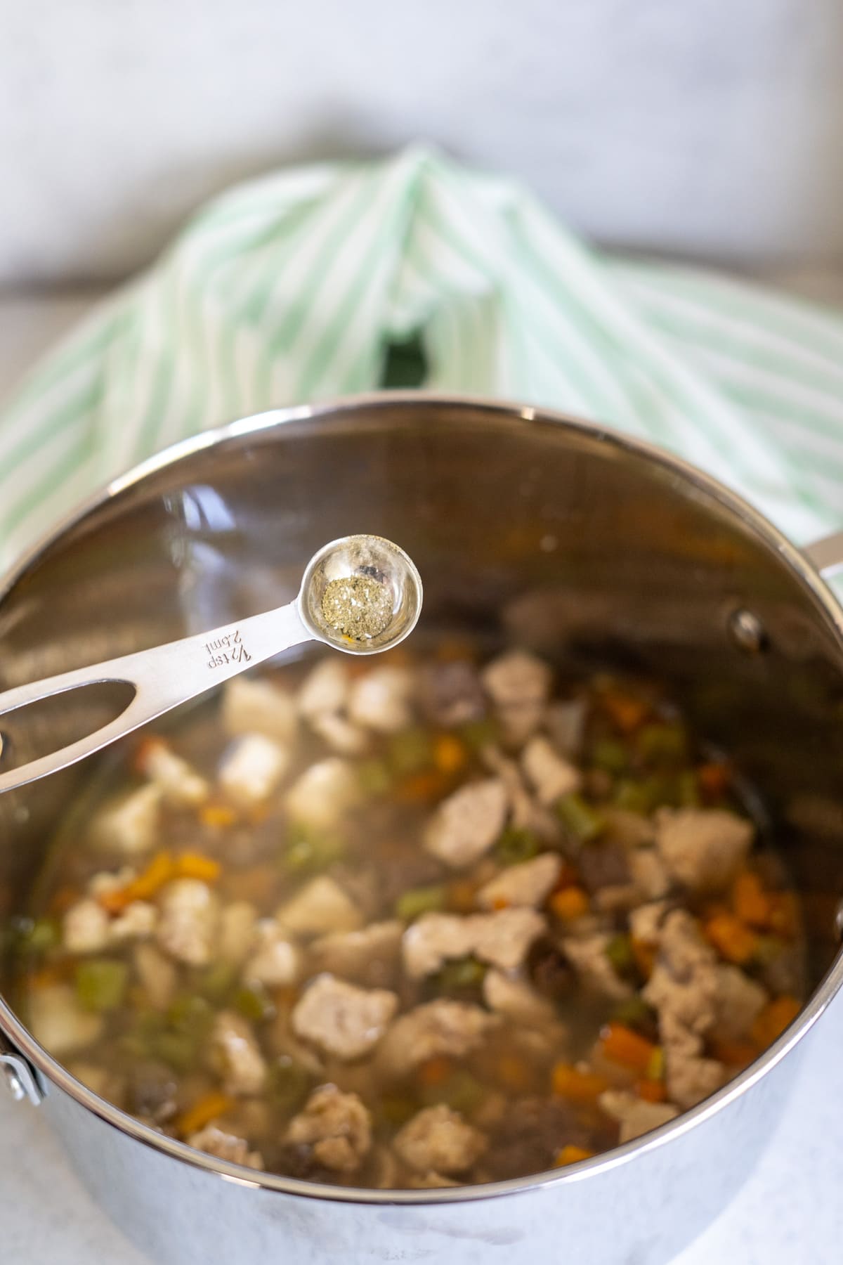 A metal saucepan filled with soup containing meat and vegetables, with a measuring spoon holding spices above it—perfect inspiration for a homemade turkey dog food recipe.
