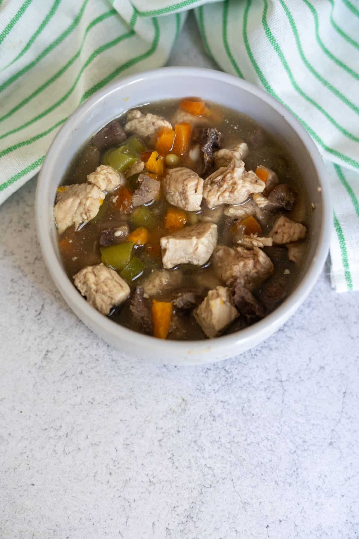 A bowl of stew with chunks of meat, diced carrots, green peppers, and mushrooms in broth—this hearty meal doubles as a wholesome turkey dog food recipe, served on a light surface next to a green-and-white striped tea towel.
