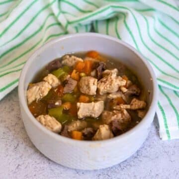 A bowl of stew containing chunks of meat and diced vegetables sits on a light surface with a green and white striped cloth in the background, resembling a wholesome turkey dog food recipe.
