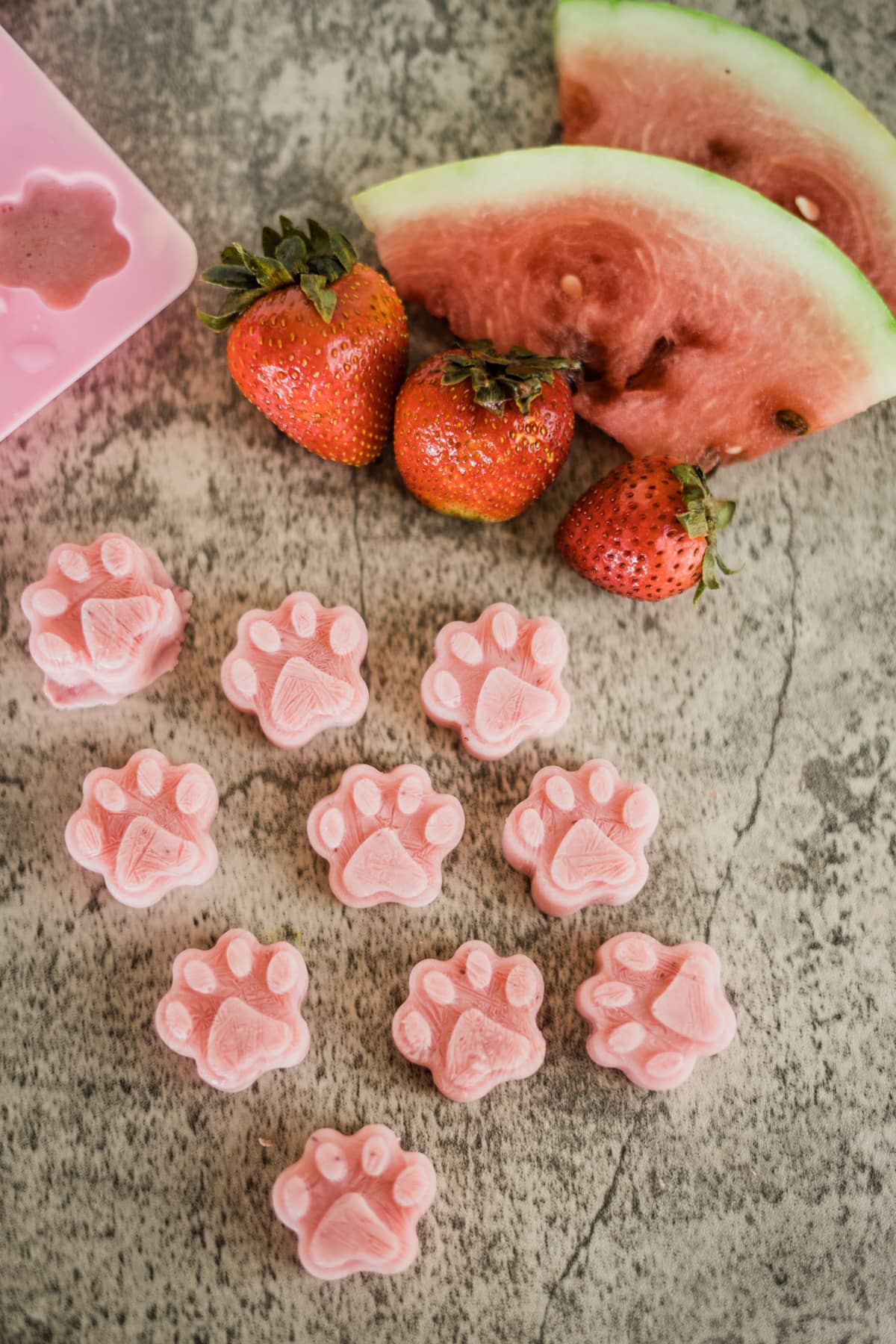 Paw shaped watermelon strawberry dog treats on a marble surface.
