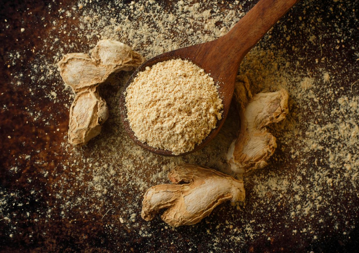 Wooden spoon with ground ginger powder rests on a dark surface, surrounded by pieces of dried ginger root and scattered powder, inviting curiosity about culinary uses—and even prompting questions like: can dogs eat ginger?.
