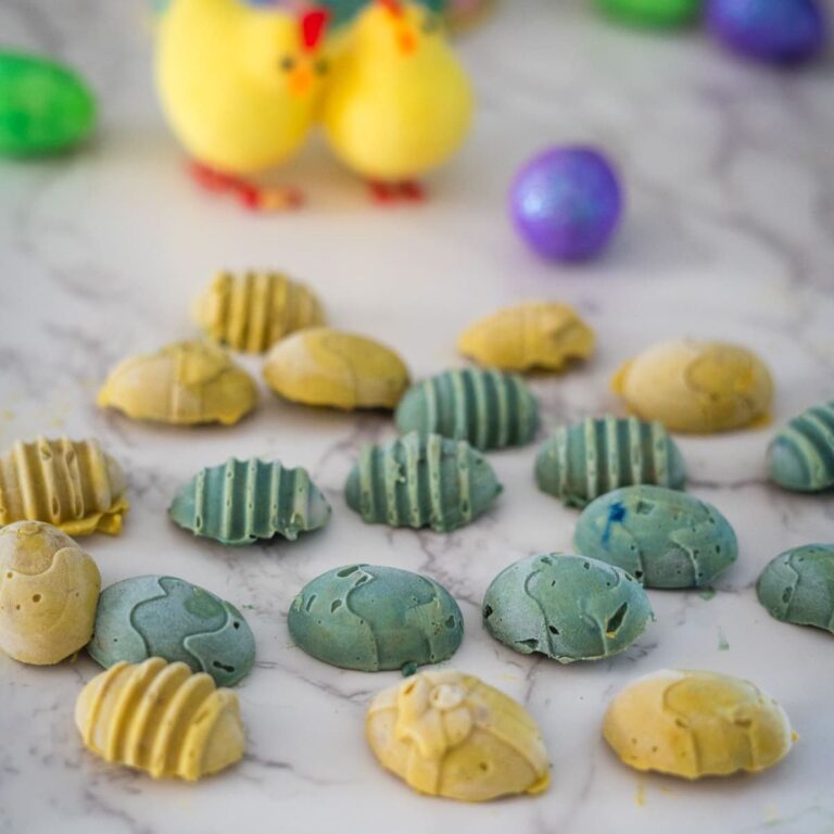 Colorful easter egg-shaped chocolates on a marble surface with decorative eggs and a yellow chick in the background.
