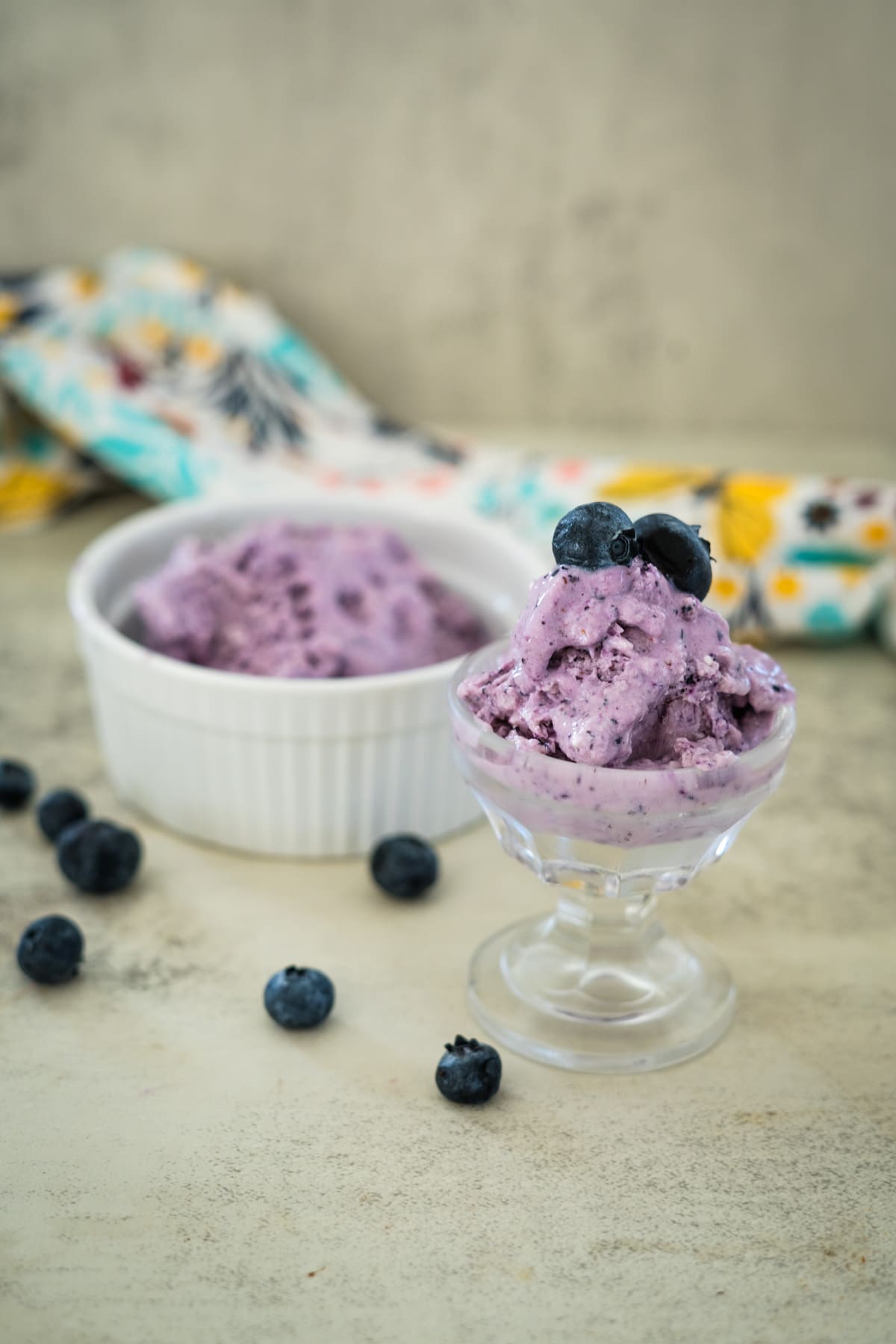 A glass dish of blueberry ice cream topped with fresh blueberries in the foreground, with another serving in a white bowl and scattered blueberries in the background on a light surface.