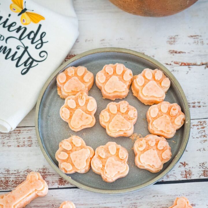 A plate with multiple paw-shaped treats is on a wooden surface next to a towel partially showing a printed word. A bone-shaped treat is also partially visible at the bottom.