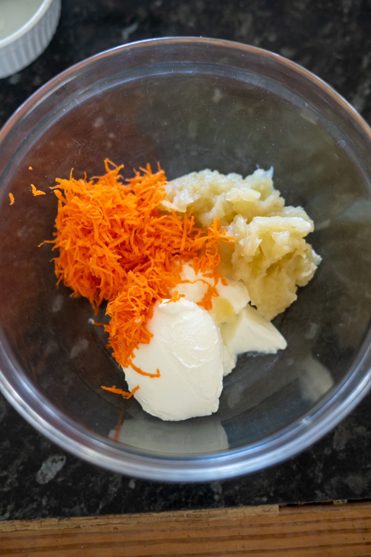 A clear bowl containing shredded carrots, mashed bananas, and a dollop of yogurt placed on a dark countertop.