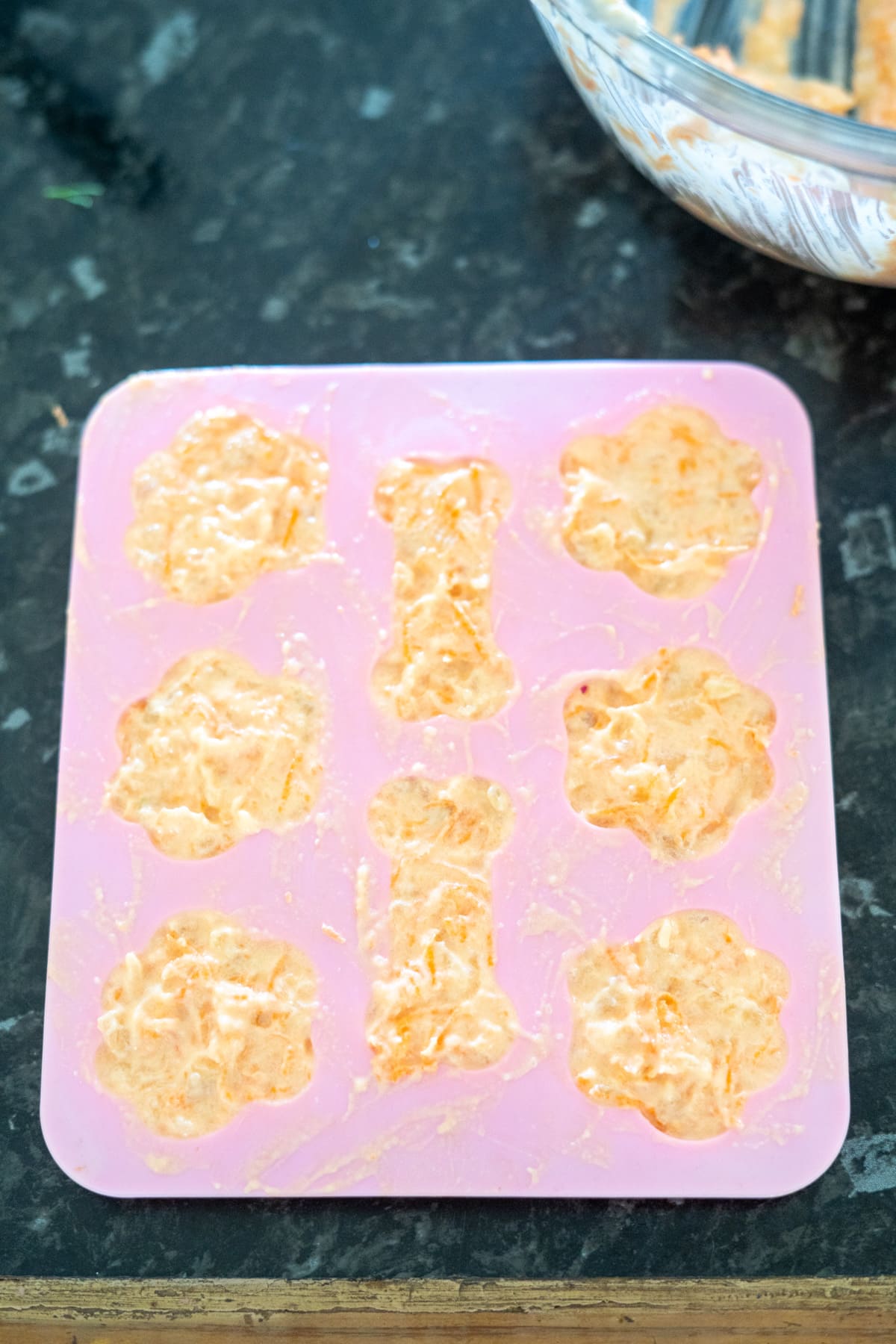 Silicone mold filled with a light orange mixture, shaped like bones and flowers, placed on a dark kitchen counter.