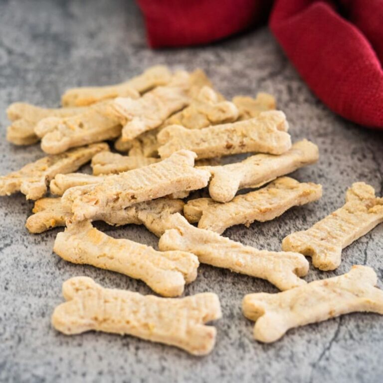 A pile of bone-shaped dog biscuits lies on a gray surface with a red cloth in the background.