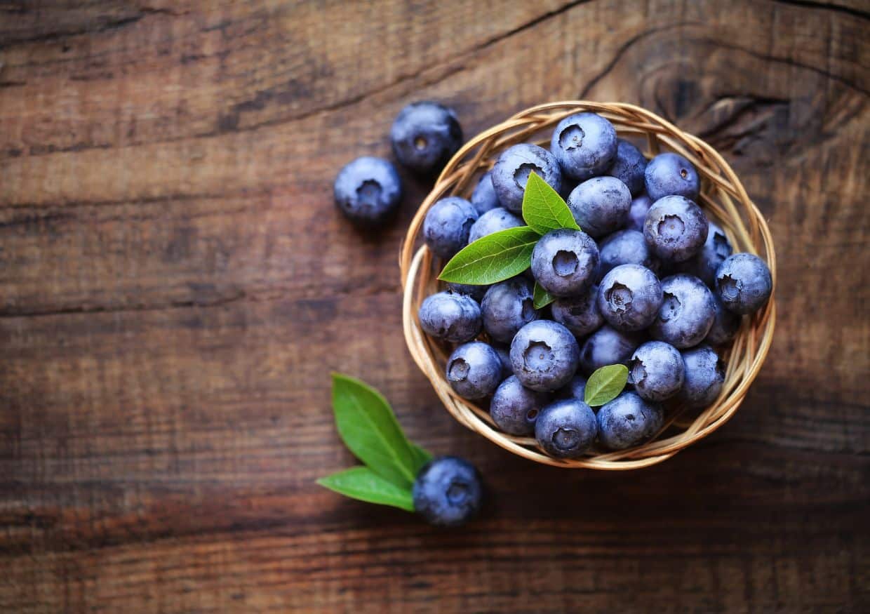 A small basket filled with fresh blueberries and a few green leaves sits on a wooden surface, perfect for inspiring blueberry dog treats recipes, with some berries and leaves scattered nearby.