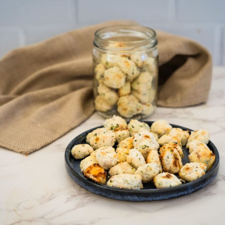 A dark plate and a glass jar filled with small, baked dumplings are placed on a marble surface with a beige cloth in the background.