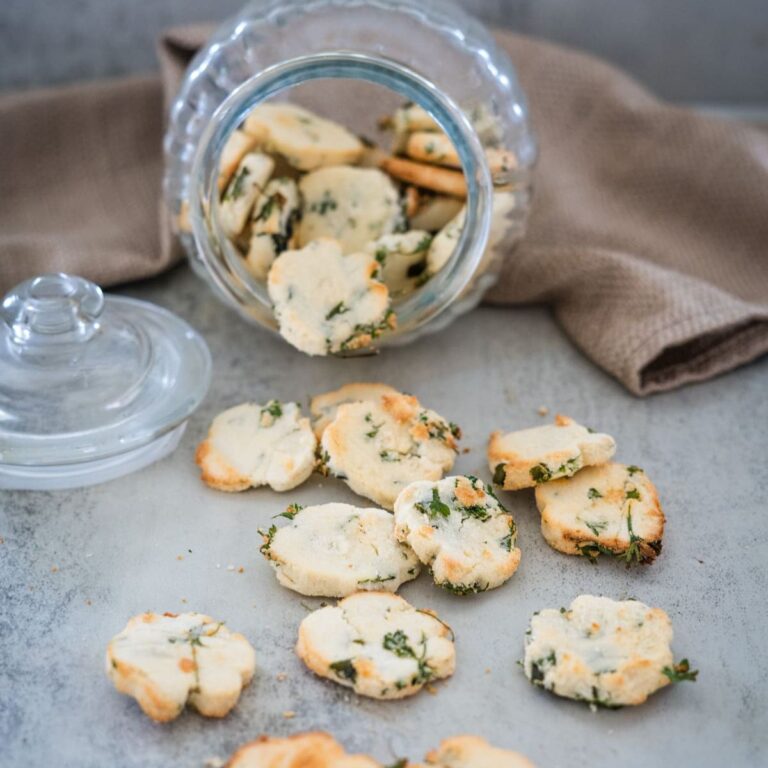 A clear glass jar is tipped over, spilling round, white cookies with small green herbs onto a gray surface, with a brown cloth in the background.
