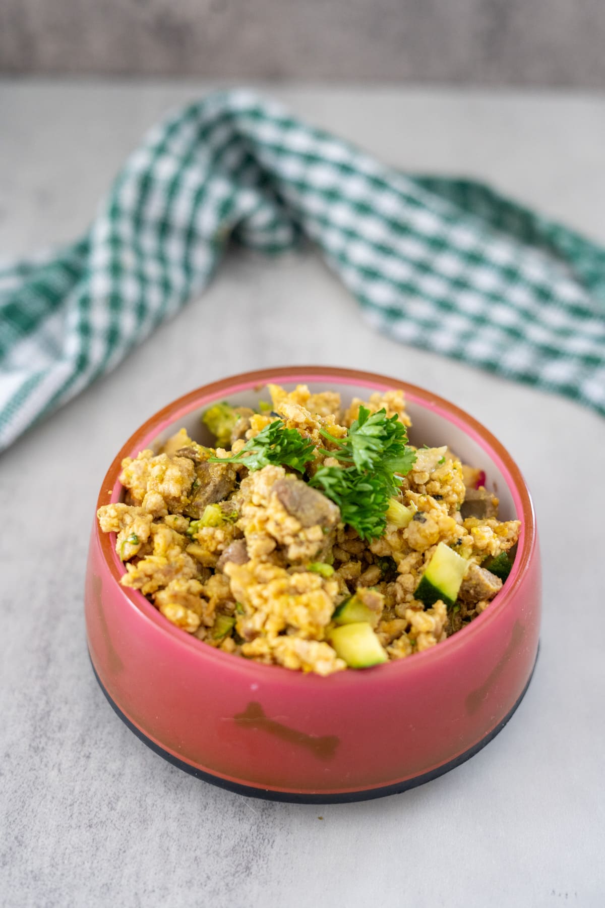 A bowl of scrambled eggs mixed with vegetables and garnished with parsley, placed on a white surface with a green and white checkered cloth in the background.