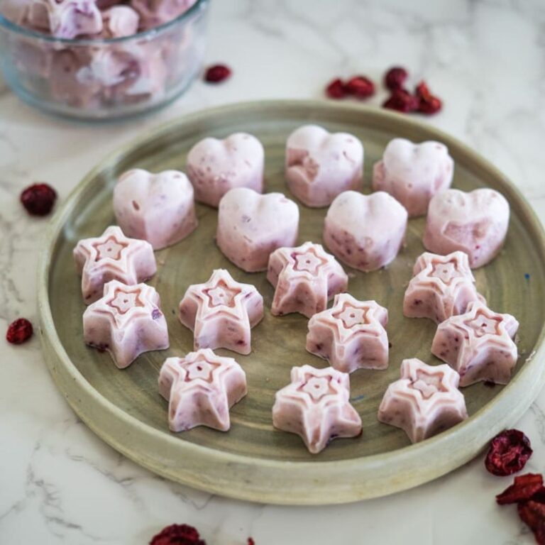 A plate of star and heart-shaped frozen yogurt bites with bits of fruit, displayed on a round gray plate with a bowl of additional bites in the background.