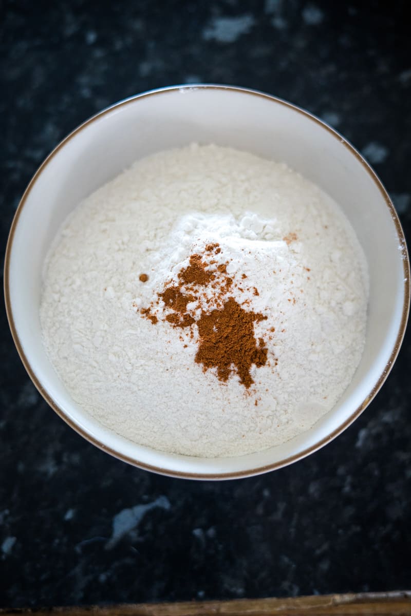 A bowl of flour with a small pile of cinnamon on top, set on a dark countertop.