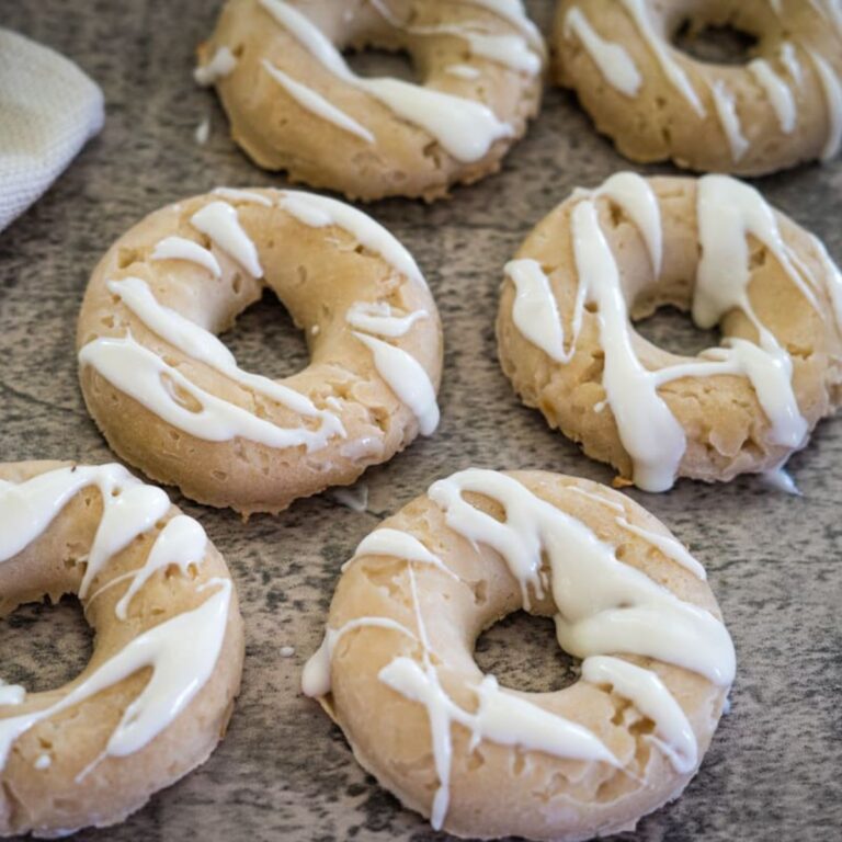 Six glazed doughnuts with white icing drizzle arranged on a textured surface.