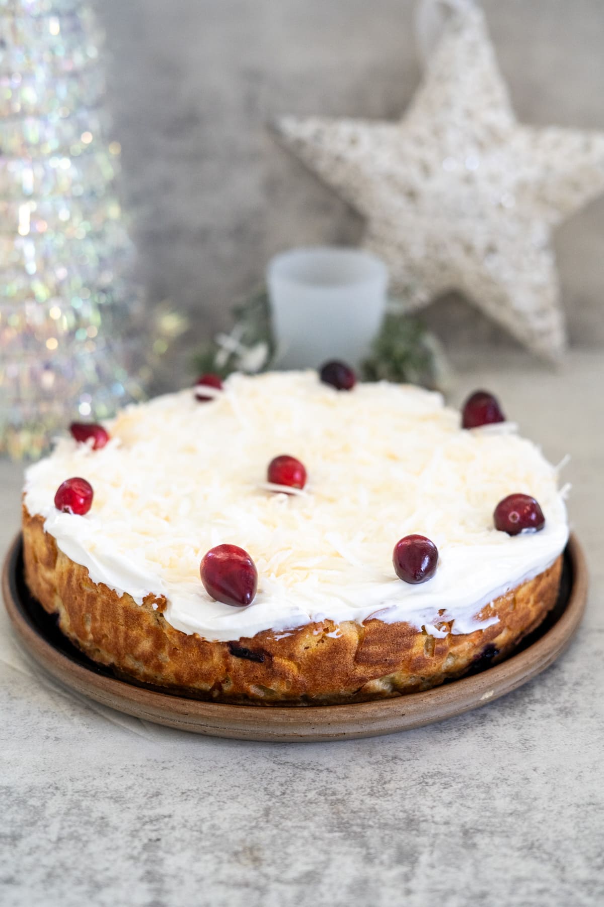 A Christmas cake with white frosting topped with cranberries is displayed on a wooden plate. A decorative star and shiny ornament, along with a cute dog figurine, add charm to the festive scene in the background.