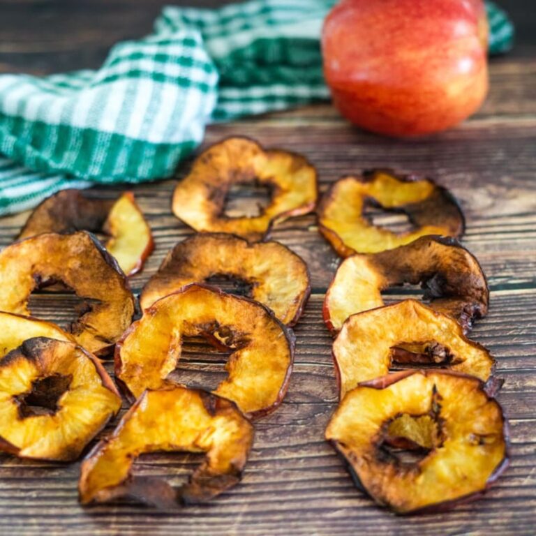 Ten slices of dried apple are arranged on a wooden surface, with a whole red apple and a green plaid cloth in the background.