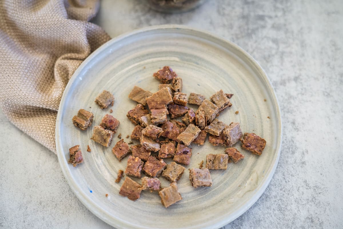 A plate with small, square, brown chicken liver cat treats rests on a light-colored, textured surface. A beige cloth is beside the plate.