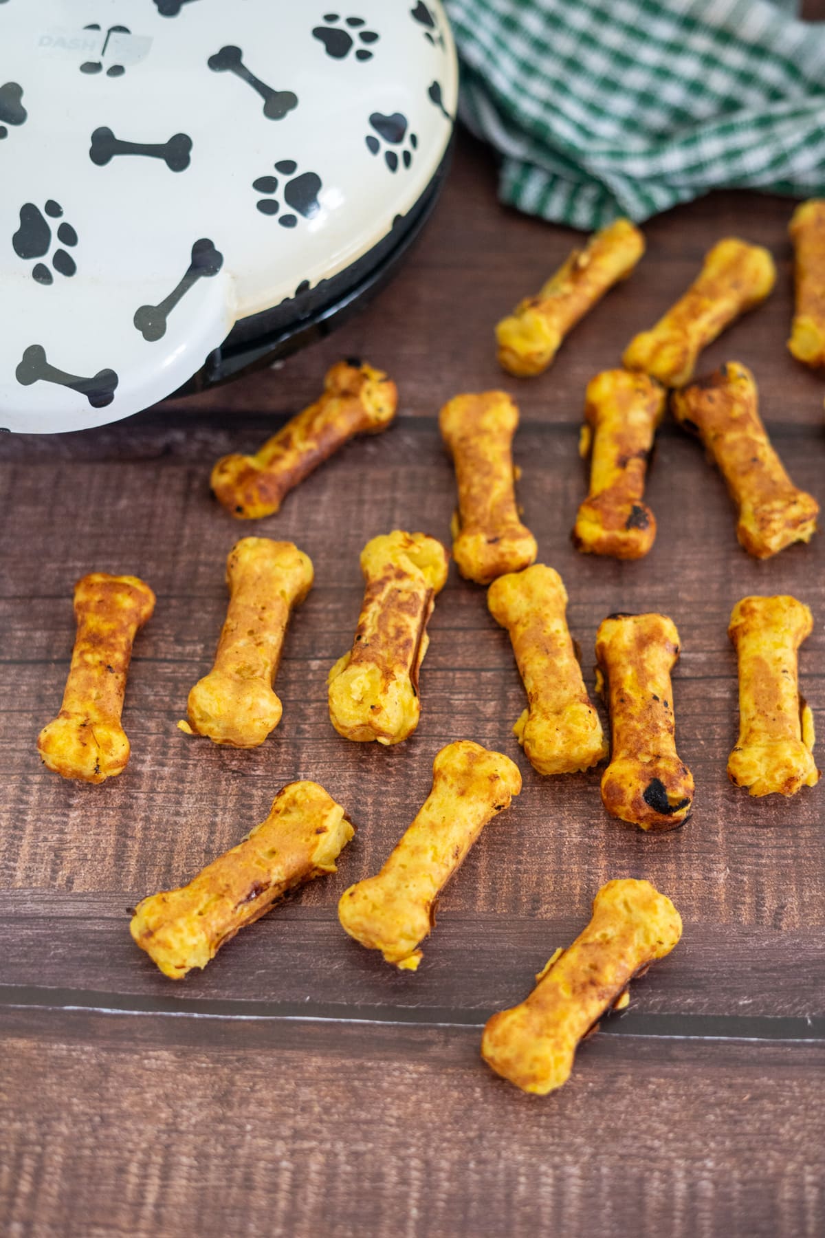 A group of pumpkin-flavored bone-shaped dog treats is scattered on a wooden surface next to a white dog bowl with black paw prints. A green checkered cloth is in the background, adding a cozy touch.