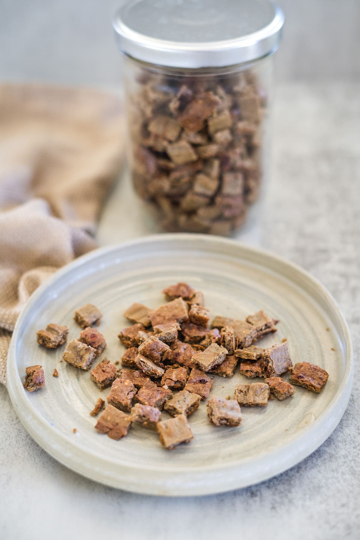 Small square dog treats arranged on a ceramic plate, with a glass jar of similar treats in the background.