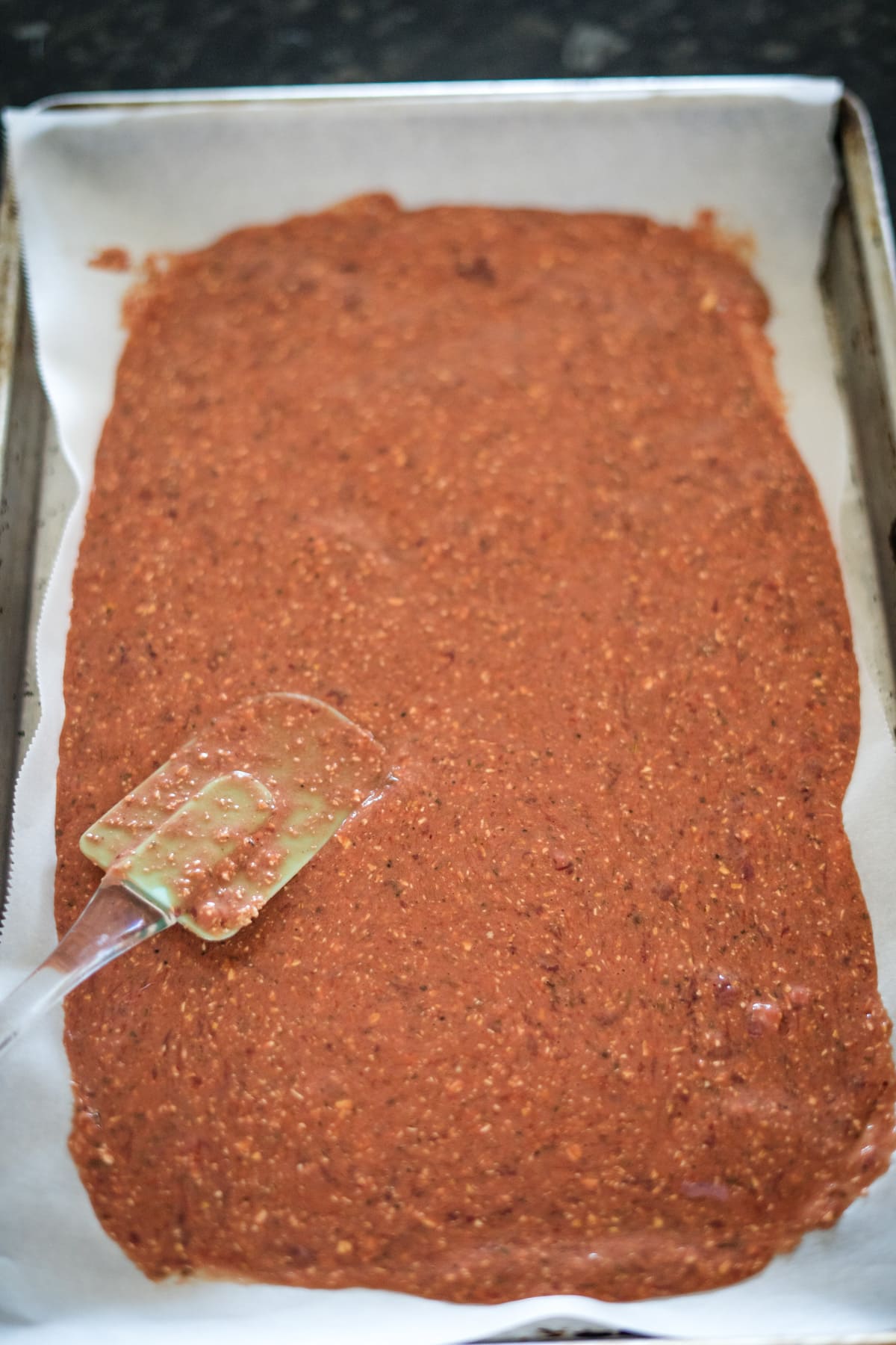 A rectangular baking sheet covered with parchment paper holds a spread of chocolate batter being smoothed by a spatula.
