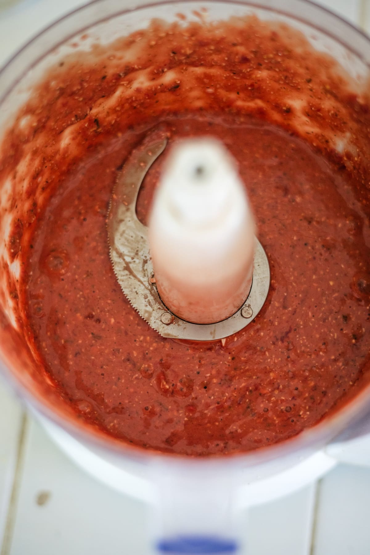Close-up of a red fruit and seed mixture in a food processor bowl with a visible blade.